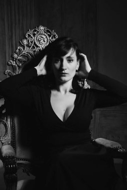 İç stüdyoda siyah seksi elbise li koltukta oturan genç güzel kadın modelsiyah beyaz portre.