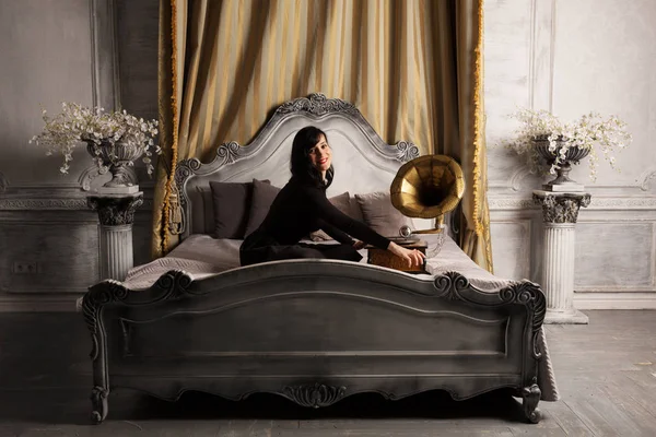 Портрет молодої красивої жінки-моделі в чорній сексуальній сукні, що сидить на ліжку з грамфонікою інтер'єру студії . — стокове фото