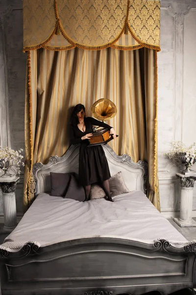 Portret van jonge mooie vrouwelijke model in zwarte sexy jurk staande op bed in met de Gramophone interieur studio. — Stockfoto