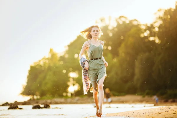 Jovem bela mulher magra com cabelo curto luz vestindo vestido de verão verde andando na costa do mar ao pôr do sol . — Fotografia de Stock