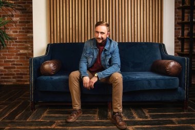 Modern apartmandaki bir kanepede oturmuş kameraya bakarken gülümseyen ve gülen mavi ceketli şık, zarif genç bir adam.