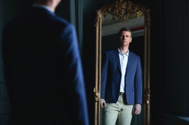 Yakışıklı, şık, uzun boylu, mavi ceketli ve bej pantolonlu adam evde aynaya bakıyor..