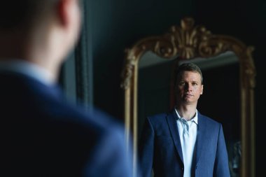 Yakışıklı, şık, uzun boylu, mavi ceketli ve bej pantolonlu adam evde aynaya bakıyor..