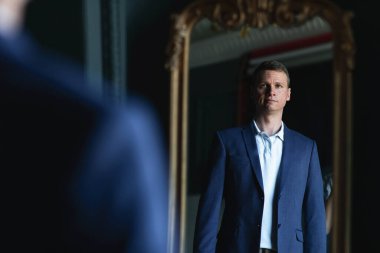 Yakışıklı, şık, uzun boylu, mavi ceketli ve bej pantolonlu adam evde aynaya bakıyor..