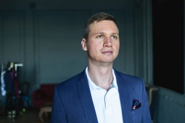 Mavi ceketli ve bej pantolonlu yakışıklı, şık bir adam. Karanlık bir odada duruyor ve klasik bir iç mimariyle yan yana bakıyor ve hayal kuruyor.