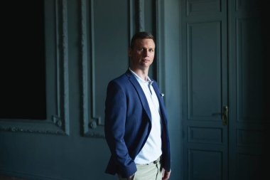 Mavi ceketli ve bej pantolonlu yakışıklı, şık bir adam. Karanlık bir odada duruyor ve kameraya bakıyor.