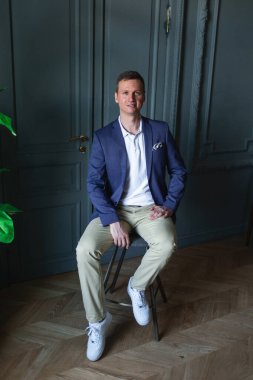 Mavi ceketli ve beyaz gömlekli yakışıklı bir adam klasik bir iç mekanı olan bir odada oturuyor, kameraya bakıyor ve gülümsüyor.