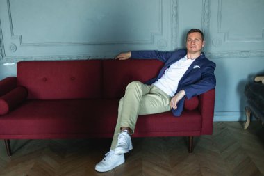 Mavi ceketli, beyaz gömlekli, şık bir adam. Kırmızı koltukta oturuyor. İçi klasik ve ciddi bir şekilde kameraya bakıyor.