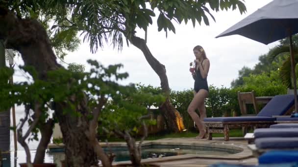 Joven americana bebiendo cóctel de frutas de pie cerca de la piscina en la isla de Bali . — Vídeos de Stock