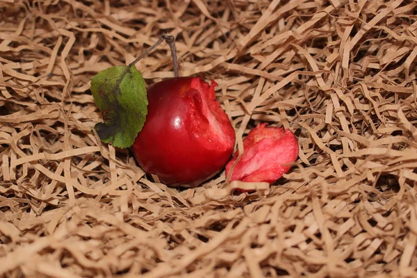 The bitten apple lies on the chopped brown paper. To the right of the apple lies a piece from it. The apple is red outside, pink inside. On the apple there is a green leaf. Front view, apple lying on its side. Close-up.