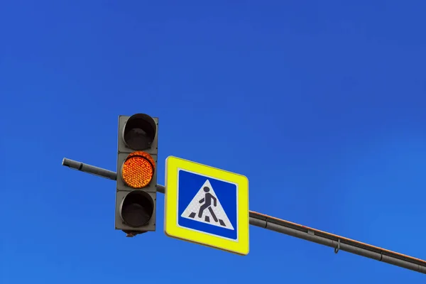 Yellow light traffic light signal and road sign of a pedestrian crossing on the console in the background of a bright sky with clouds. Copy space for text