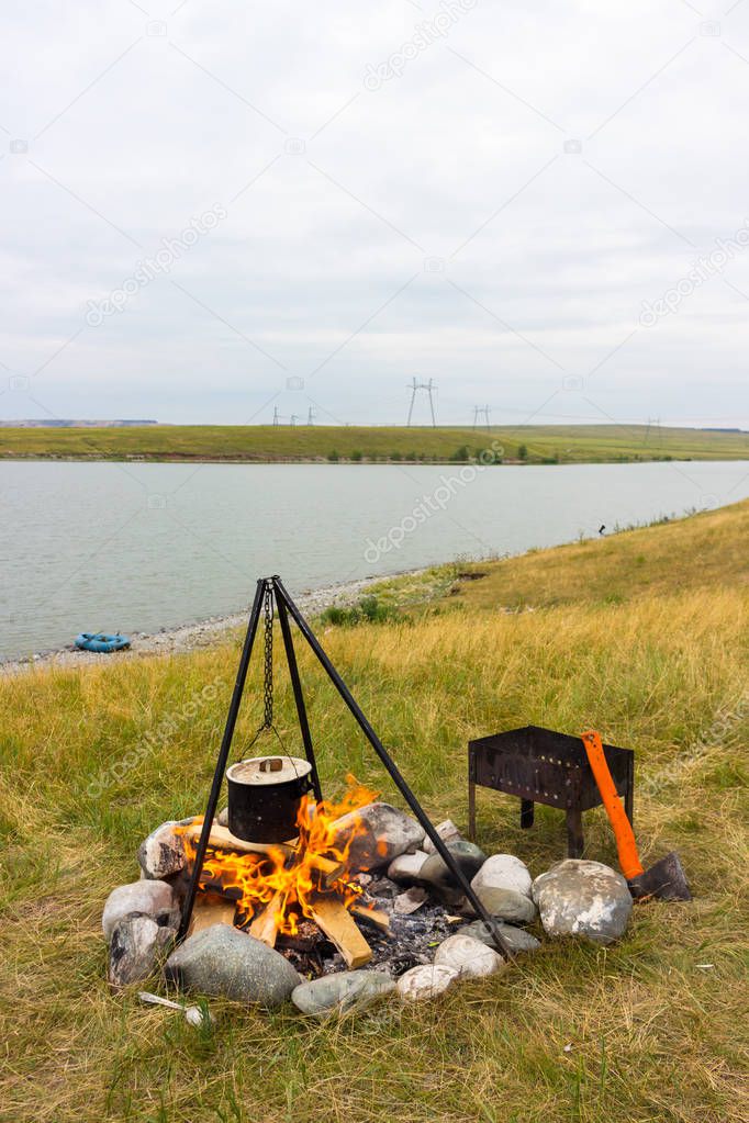 Cooking in black sooty saucepan on bonfire on the shore lake. Authentic scene from travel in time summer vacation. Soup on nature