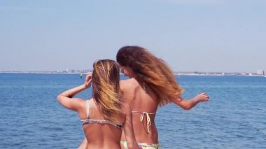 İki genç güzel kız bikini yürüyerek deniz arka plan üzerinde bir iskelede. Ağır çekim.