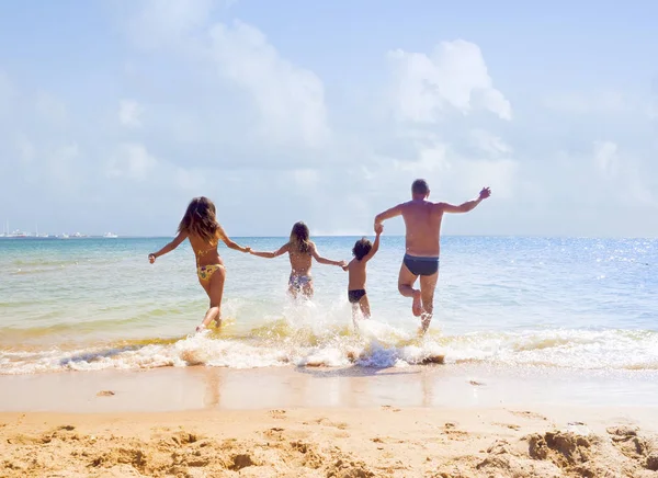 Una familia feliz de madre, padre y dos hijos, hijo e hija, corriendo al mar tomados de la mano y divirtiéndose en la arena de una playa soleada — Foto de Stock