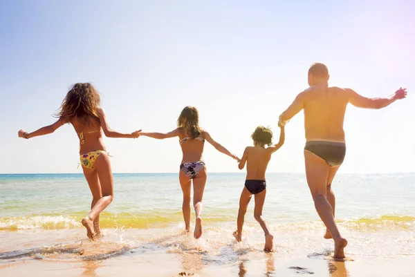ครอบครัวที่มีความสุขของแม่, พ่อและลูกสองคน, ลูกชายและลูกสาว, วิ่งเข้าไปในทะเลจับมือกันและมีความสุขในทรายของชายหาดที่มีแดด — ภาพถ่ายสต็อก