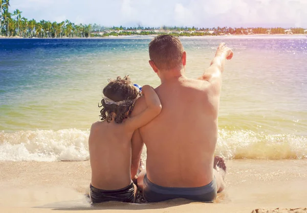 Padre e hijo se divierten en la playa tropical de arena blanca. Vista trasera del padre y el hijo sentados en la arena mirando al océano — Foto de Stock
