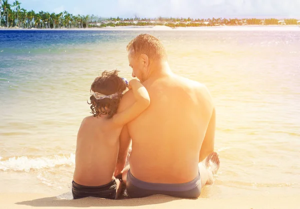 Padre e hijo se divierten en la playa tropical de arena blanca. Vista trasera del padre y el hijo sentados en la arena mirando al océano — Foto de Stock