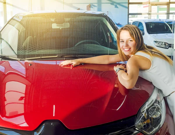 Dream about car. Gorgeous smiling woman hugging lies on the hood of new red car in the dealership.