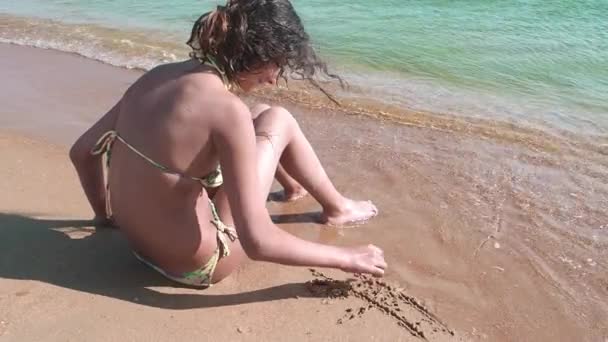 可爱的女孩在沙滩上画了一个脚趾 海浪冲刷着图案 慢动作 — 图库视频影像