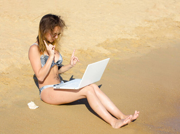 A student sitting on the coast with a laptop and raising her finger upwards in a surprised and shocked expression. Girl with laptop on tropical beach. remote working concept.