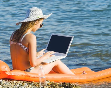 Tropik sahilde laptop ile çalışan güzel genç kadın. Hasır şapka sarı şişme yatak üzerinde oturan tatillerde dizüstü kullanarak mutlu seyahat kadın.