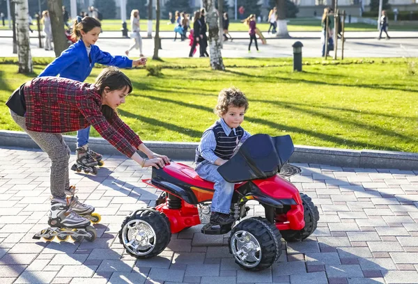 Kids driving electric toy car in summer park. Children in battery power vehicle. A girl riding on roller skates clinging to the car. Little boy and his two older sisters have fun on the city street. — Stock Photo, Image