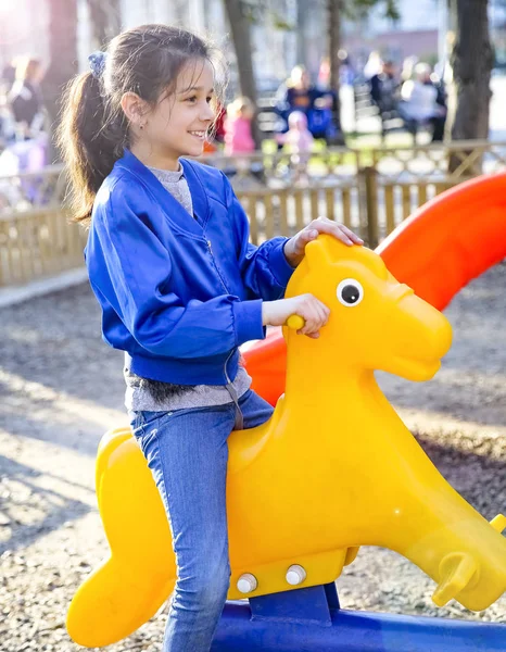 Little girl swinging on a plastic yellow horse. The concept of a happy childhood, games in the city park, in kindergarten and in the family. — Stock Photo, Image