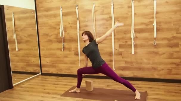 Deportiva hermosa madura principiante estudiante de yoga de pie en Utthita Trikonasana, Triángulo Extendido Pose apoyada en ladrillo de madera yóguica, vista de perfil sobre un fondo de madera, cámara lenta — Vídeo de stock