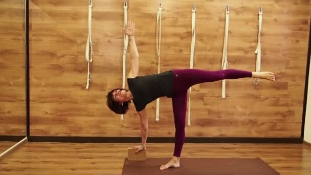 Sportif belle mature débutant yoga étudiant femme debout dans Utthita Trikonasana, Extended Triangle Pose appuyé sur la brique de bois yogique, vue de profil sur un fond en bois, ralenti — Video