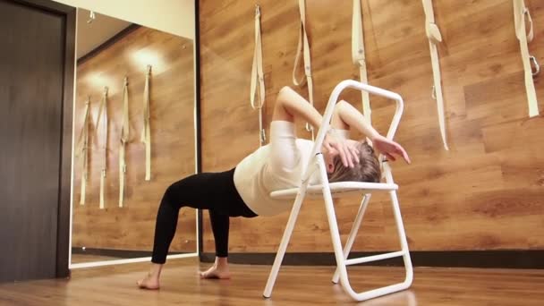 Profesora de Yoga practicando en estudio con paredes de madera y suelo. Yogui usando silla para la pose parsvottanasana. Instructor de yoga Iyengar con silla como puntal para ayudar en la postura, cámara lenta — Vídeo de stock