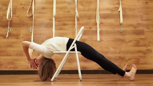 Учитель йоги практикует в студии с деревянными стенами и полом. Йоги использует стул для позирования parsvottanasana. Инструктор йоги Iyengar со стулом в качестве опоры, чтобы помочь в осанке, замедленной съемки — стоковое видео