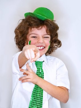 Kıvırcık çocuk çocuk eğleniyor Saint Patrick kutlamalar sırasında hafif bir arka plan üzerinde. Kameraya bir parmak işaret yanağımda bir İrlanda bayrağı çocukla gülümsüyorum