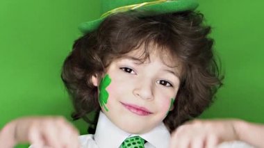 Çocuk kutlayan St. Patrick's günü onun makyaj gösterilen. Küçük, düzgün vücutlu erkek Yaprak yonca ve sevgi jesti gösterilen yanakları İrlanda bayrağı. ağır çekim. yeşil arka plan