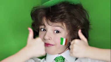 Çocuk kutlayan St. Patrick's günü onun makyaj gösterilen. Yaprak yonca ve İrlanda bayrağı bir jest iyi gösterilen yanakları üzerinde küçük, düzgün vücutlu çocukla. ağır çekim. yeşil arka plan