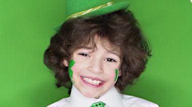 Çocuk kutlayan St. Patrick's gün kameraya bakarak. Bir yaprak yonca ve İrlanda bayrağı şeklinde yanaklarını üzerinde çizim ile bir küçük, düzgün vücutlu erkek yüz eller geçerlidir. ağır çekim.