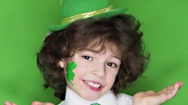 Çocuk kutlayan St. Patrick's gün kameraya bakarak. Bir yaprak yonca ve İrlanda bayrağı şeklinde yanaklarını üzerinde çizim ile küçük, düzgün vücutlu bir çocuk. ağır çekim. yeşil arka plan
