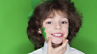 Çocuk kutlayan St. Patrick's günü onun makyaj gösterilen. Bir küçük, düzgün vücutlu erkek onun yanağına bir yaprak yonca ve İrlanda bayrağı şeklinde bir çizim, onun parmak işaret eder. ağır çekim. yeşil arka plan