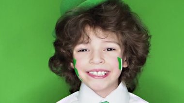 Çocuk kutlayan St. Patrick's gün kameraya bakarak. Bir yaprak yonca ve İrlanda bayrağı şeklinde yanaklarını üzerinde çizim ile küçük, düzgün vücutlu bir çocuk. ağır çekim. yeşil arka plan