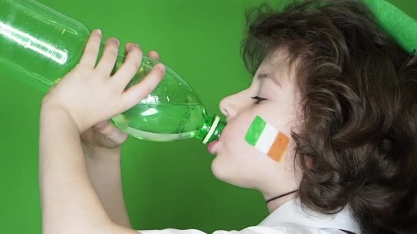 帽子と彼の頬にアイルランドの旗のイメージと幸せな少年は 緑の背景に分離されたボトルから水を飲みます プロファイル スローモーション — ストック動画