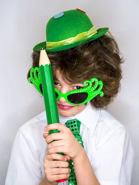 Komik St Patricks günü yeşil bir şapka, karnaval gözlük ve bir kravat giyen küçük çocuk çarpıcı. Kıvırcık çocuk büyük yeşil kalem hafif bir arka plan üzerinde el ile poz