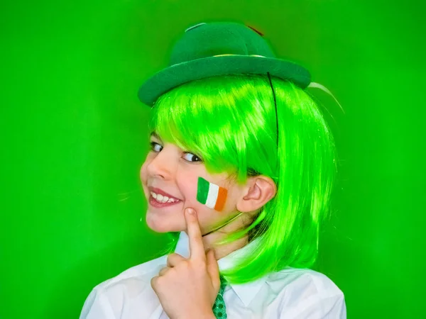 庆祝圣帕特里克节的孩子展示他的化妆 一个穿着绿色狂欢节配饰的卷曲的小男孩指着他脸颊上的爱尔兰国旗 绿色背景 — 图库照片