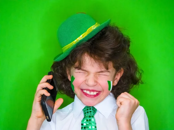有趣的圣帕特里克日惊人的小男孩戴着绿色的帽子和洗发水 他脸颊上的爱尔兰国旗在电话里说话 — 图库照片