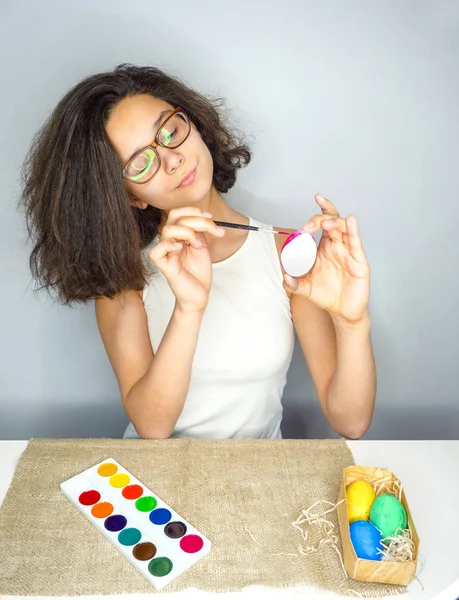 The girl to paint eggs to the Christian holiday.Happy pretty girl is preparing for the celebration of Easter, painting eggs, sitting at the table.Gray background.