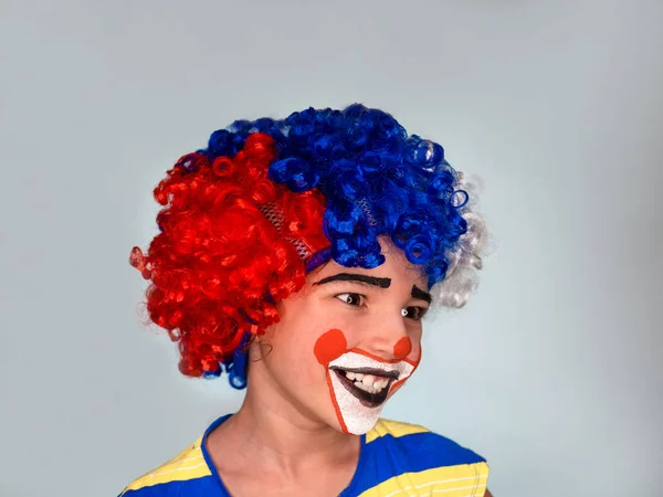 Kleine schattige lachende jongen in een kleurrijke pruik met facepaint als clown kijkt weg, pantomime, emoties. April Fool's Day, April 1. geïsoleerd op grijs. — Stockfoto