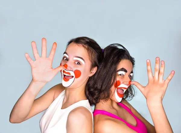 Dos morenas adolescentes en camisetas y con caras pintadas bromeando muestran sus largas narices con la palma de sus dedos. Aislado en gris. La familia celebra el Día de los Inocentes . — Foto de Stock