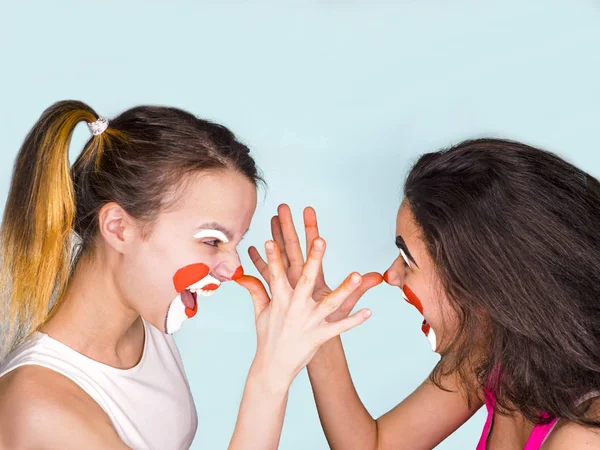 Dos morenas adolescentes en camisetas y con caras pintadas bromeando muestran sus largas narices con la palma de sus dedos. Aislado en gris. La familia celebra el Día de los Inocentes . —  Fotos de Stock