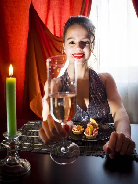 Sensual girl holding a glass of champagne, drinking champagne in a restaurant, in an evening shiny dress, on a date or a meeting, looking into the camera and tries to touch it. Selective focus