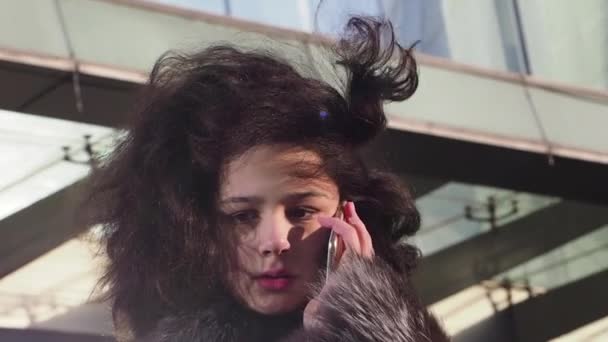 昼間にスマートフォンで話す冬服の若いカーリーの女の子 近代的な都市の背景 スローモーション — ストック動画