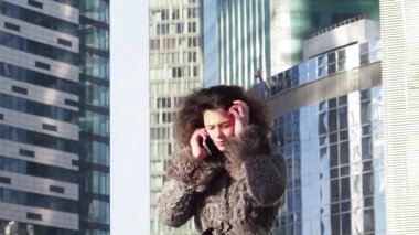 Kış giysileri genç kıvırcık kız gündüz akıllı telefon konuşurken, modern şehir arka plan. Yavaş çekim 