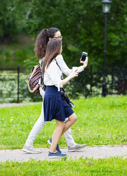 Two friends having walk in park in Russian traditional park, Moscow. Young girls communicating and using mobile phones by nature pool. Young women travelers browse map app